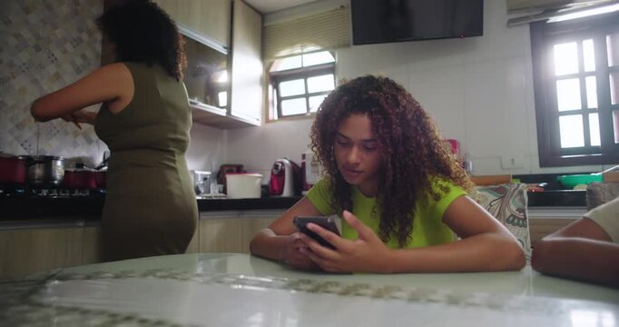 Teenage girl smiling while looking at phone, enjoying content at dining table, mother cooking in kitchen in background, joyful family moment, family of African descent