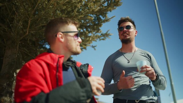 Two male friends enjoy warm drinks and conversation on a sunny winter day. Young men share a friendly moment outdoors, sipping beverages under the clear sky.