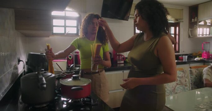 Family cooking together in kitchen, mother stirring pot, daughter adding pasta, father drinking coffee, young boy working on laptop at table, family of African descent