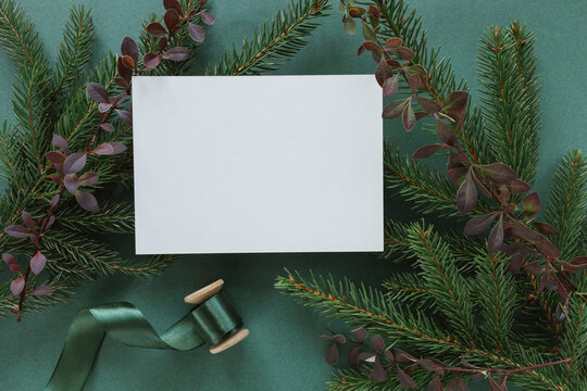 Overhead view of a blank card on evergreen fir branches, barberry sprigs and a spool of ribbon on a green background