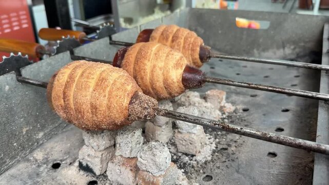 Trdelnik Spiral pastries are being baked on skewers over hot coals, revealing a golden-brown crust in a vibrant street food environment