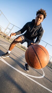 Vertical photo of a young Black man dribbling a basketball on an outdoor court. Low angle wide angle shot with perspective distortion and dutch tilt. Athletic streetball player training