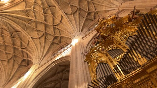 Low angle panning shot of the ornate golden pipe organ and intricate vaulted ceiling inside the historic Segovia Cathedral in Segovia, Spain.