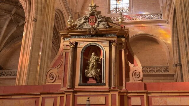 Slow zoom shot of an ornate religious statue set within an intricate marble altarpiece under colorful stained glass light inside the historic Segovia Cathedral in Spain.
