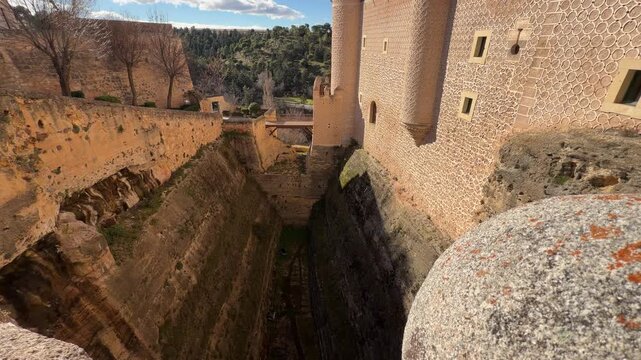 High angle perspective shot overlooking the deep moat and historic stone walls of the Alcazar castle, a monumental medieval fortress under a clear sky in Segovia, Spain.