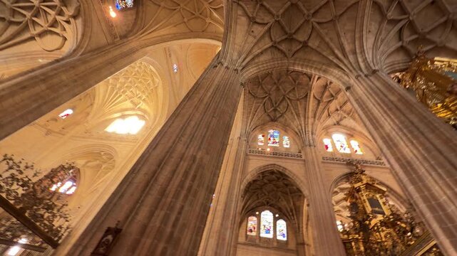 Low angle panning shot of the majestic vaulted ceilings, intricate stone pillars, and vibrant stained glass windows inside the historic Segovia Cathedral in Segovia, Spain.