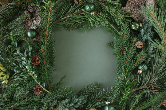 Overhead flatlay of assorted winter foliage including evergreen branches, pinecones, tree bark and Christmas baubles arranged as a border on a green background