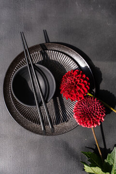 Close-up overhead view of an Asian place setting with a bowls, chopsticks and two dark red Pompon Dahlias on a grey concrete table