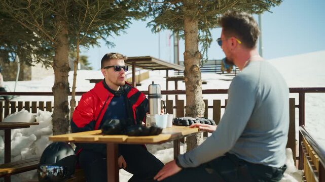 Two friends take a break from skiing to enjoy a warm drink at an outdoor cafe in the snowy mountains.