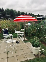 A paved garden patio features white folding furniture under a red and white striped parasol in a blooming garden. © Michael