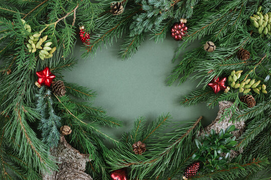 Overhead view of assorted fir branches, thuja branches, pinecones and red Christmas ornaments arranged in a border on a green background