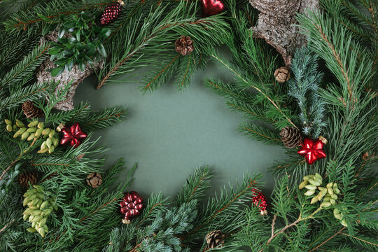 Overhead view of evergreen fir branches, thuja, pinecones and assorted festive red Christmas baubles and ornaments arranged as a border on a green background