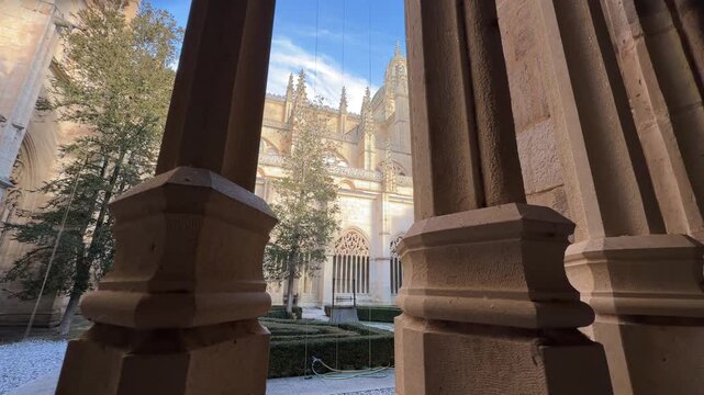 Low angle frame through ornate stone columns revealing the gothic architecture and sunlit cloister garden of the historic Segovia Cathedral in Spain.