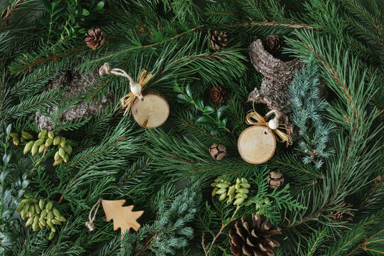 Full frame overhead flatlay of assorted evergreen foliage including fir branches, thuja, tree bark and pinecones with various wooden Christmas ornaments