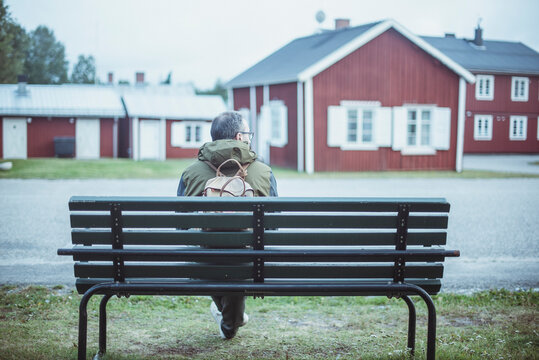 Rear view of a Man wearing a rucksack sitting on a roadside bench in front of traditional houses, Sweden