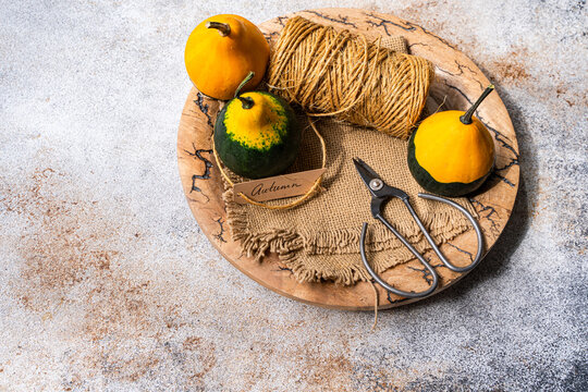 Overhead view of three decorative mini pumpkins, a ball of string, scissors, burlap fabric and a gift tag with the word autumn on a Wooden board