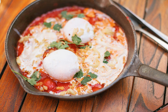 Close-up overhead view of a cast iron frying pan with tomato, poached eggs, stracciatella cheese and cilantro (shakshuka) on a wooden table