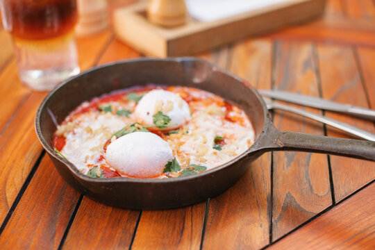 Close-up of a cast iron frying pan with tomato, poached eggs, stracciatella cheese and cilantro (shakshuka) on a wooden table with an iced espresso tonic coffee