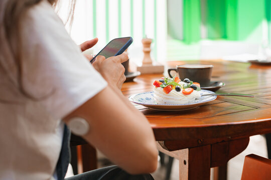 Close-up rear view of a Diabetic woman sitting at a table checking blood sugar levels on a  smartphone app before eating a meringue fruit dessert (pavlova)