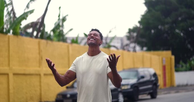 Man standing with arms raised in spiritual gratitude and devotion, head tilted back, expressing joy and connection, urban neighborhood blurred in background