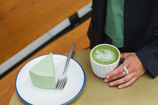 Close-up of a woman sitting an outdoor cafe table with a hot matcha latte and slice of green matcha mousse cake