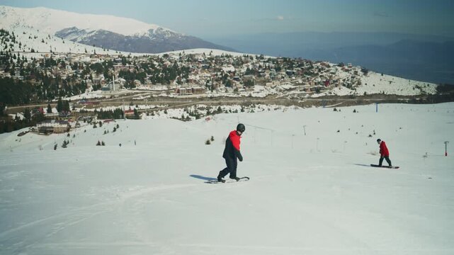 Two snowboarders descend a snow-covered mountain slope on a bright winter day. The scenic ski resort village and distant snowy peaks create a beautiful backdrop.