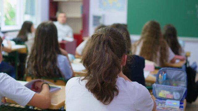 A group of young students sits at wooden desks in a bright classroom, while a classmate writes lesson notes on the large green chalkboard at the front of the room