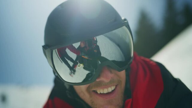 A man wearing a ski helmet and goggles smiles on a sunny winter day. His goggles reflect friends skiing down the snowy mountain.