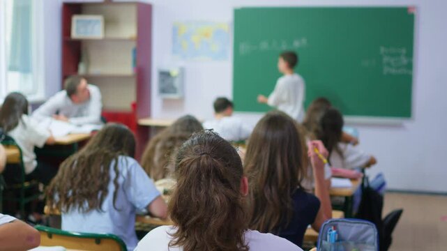 A group of young students sits at wooden desks in a bright classroom, while a classmate writes lesson notes on the large green chalkboard at the front of the room