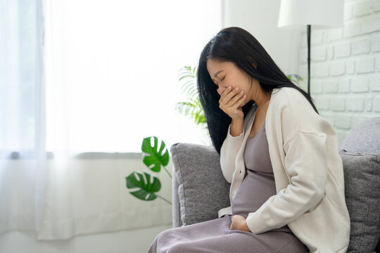 Pregnant Asian woman covers her mouth with her hand and suffers from morning sickness while sitting on the sofa in the living room.
