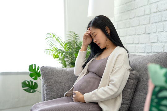 Asian pregnant woman experiencing gestational depression sitting on a sofa in the living room. Family planning concept.