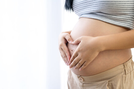 Pregnant Asian woman is making a heart shape with her hands on her large belly. She is standing by a window at home. Concepts related to pregnancy and childbirth.