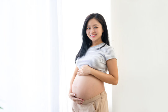 Portrait of young pregnant woman standing near window holding hands on her belly at modern home