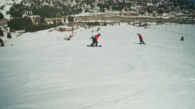 Two snowboarders are seen descending a snow-covered mountain slope, enjoying their winter sport. A scenic view of a ski resort town and distant peaks completes the picturesque scene.