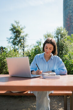 Student Taking Notes in Planner Beside Laptop in Sunny Park