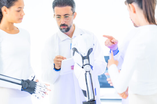 Artificial intelligence research team at a benchtop robot arm: a colleague points out a control detail while the lead engineer adjusts the manipulator in a white lab