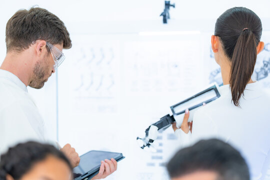 Haptic feedback engineering review at a wall of robot-hand flexion schematics, where an engineer takes notes on a tablet while a colleague holds up a black robotic arm