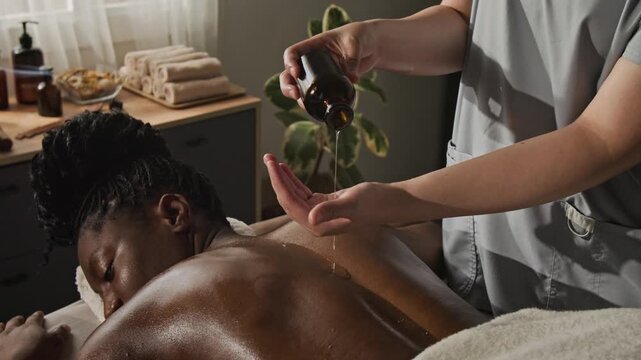 Caucasian female masseuse pouring essential oil onto young adult African American woman back. Ideal for spa salon advertising, body care promotions, and wellness marketing
