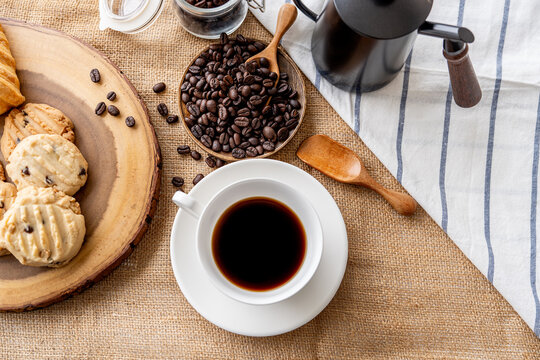 A white cup of dark coffee is arranged with coffee beans, a vintage grinder, and bread rolls on a natural tabletop.
