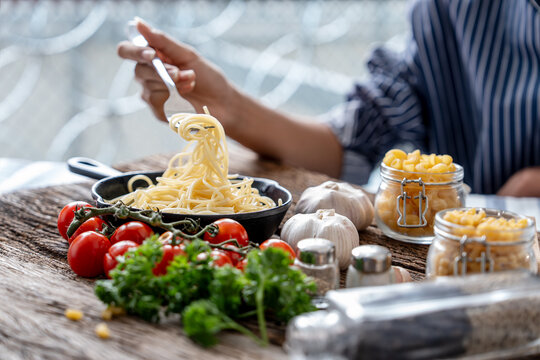 A person twirls cooked spaghetti with a fork above a black skillet surrounded by tomatoes garlic parsley and dry pasta on a rustic wooden table.