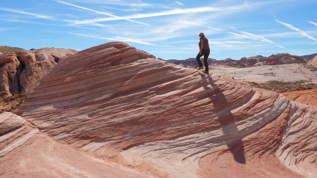 Woman trekking on colorful striped sandstone fire wave, navigating unique geological formation in valley fire state park, nevada. Tourist climbs up colorful monolithic sandstone with wave like pattern