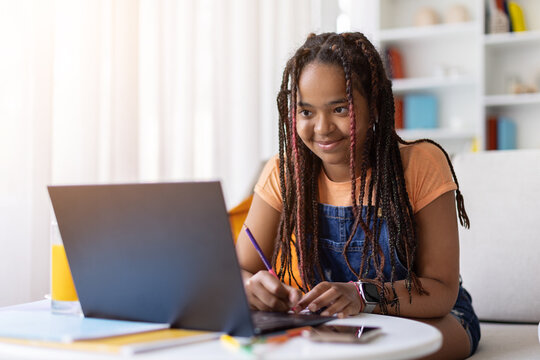 Black teenage girl studying at home with laptop, writing notes with pencil while smiling at the screen, surrounded by notebooks, school supplies, juice and a smartphone