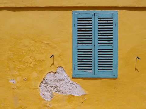 Vue rapproch&eacute;e d'une fen&ecirc;tre rectangulaire ferm&eacute;e aux volets en bois bleus, au sein d'un mur ancien peint en jaune et pr&eacute;sentant une tache blanche.