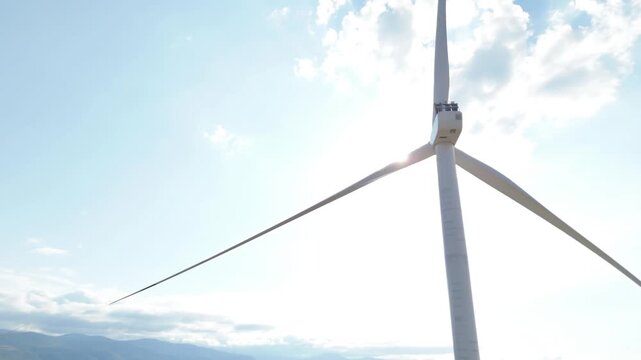 Close up of a large wind turbine rotating slowly, generating clean and renewable electricity with the bright sun flaring behind its blades against a beautiful blue sky