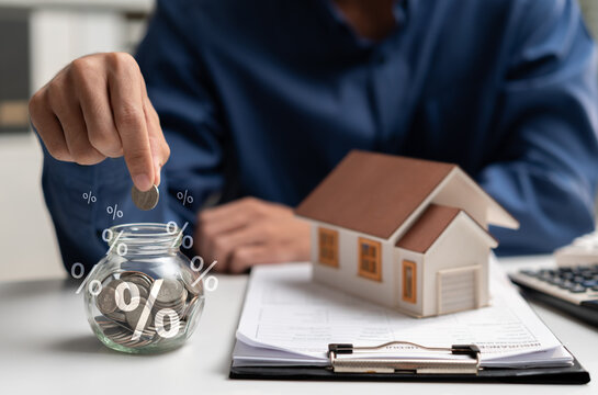 Home finance concept with person saving coins beside a house model and percentage symbols, representing mortgage interest, down payment, property investment, loan planning, and household budgeting.