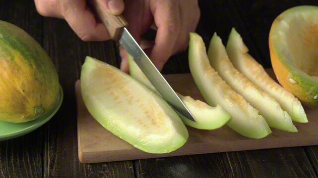 Hand slicing green melon fruit on wooden cutting board with knife, showcasing continuous action of cutting, revealing juicy flesh, and displaying whole melon and slices on rustic table