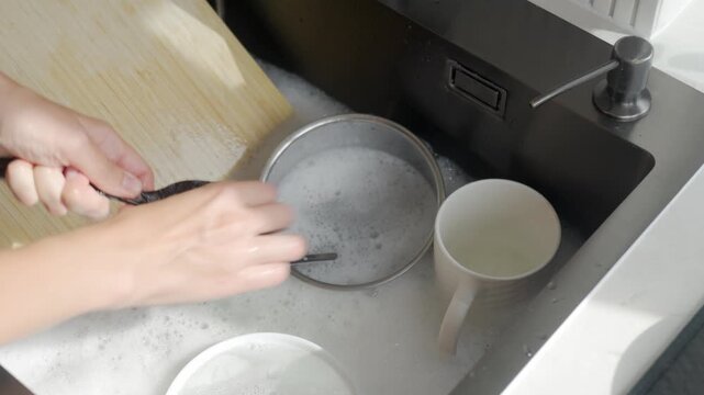 Woman cleans black cooking spoon with sponge over soapy kitchen sink while other dishes soak nearby. Washing kitchen utensils and cleaning up after cooking