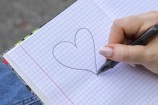 Dłoń rysująca serce czarnym długopisem na papierze w kratkę, minimalistyczny symbol miłości, uczuć i romantycznego wyznania.