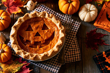 Fototapeta na wymiar Pumpkin pie with jack-o-lantern face cut into crust, Halloween overhead flat lay with pumpkins and gingham napkin