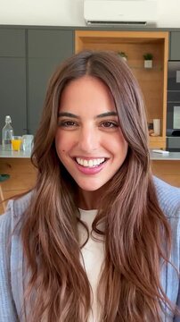 A beautiful young woman with long brown hair smiles and laughs directly at the camera in a modern home setting, conveying happiness, joy, and positive emotion.